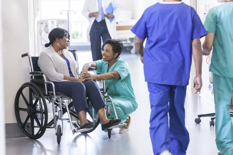 Caring African American female nurse in hospital uniform scrubs with wheelchair patient in medical center